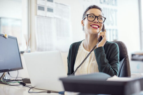 Smiling young businesswoman talking on landline phone. Female professional is using telephone while looking away. She is working at desk in creative office.